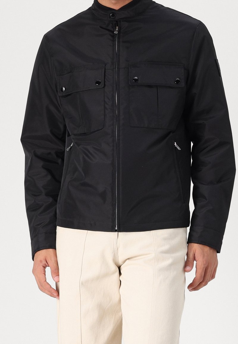 Černá bunda na zip s vysokým límcem, vybavená dvěma náprsními kapsami a bočními kapsami na zip. Vyrobena z hladké tkaniny s matným povrchem.