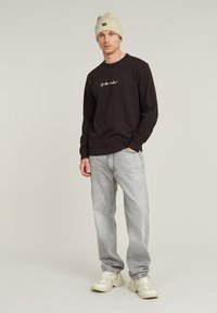 Dark brown sweatshirt with logo detail, gray jeans, and cream beanie. Model wears beige sneakers, standing in a neutral background.