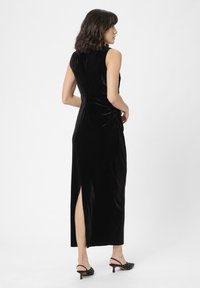 Donna che indossa un abito midi in velluto nero senza maniche con arricciature laterali e uno spacco posteriore, abbinato a scarpe con tacco slingback nere.