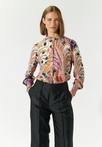 Blusa multicolore con motivo floreale e a spirale, con bottoni davanti, collo alto e maniche con polsini, abbinata a pantaloni neri a vita alta.