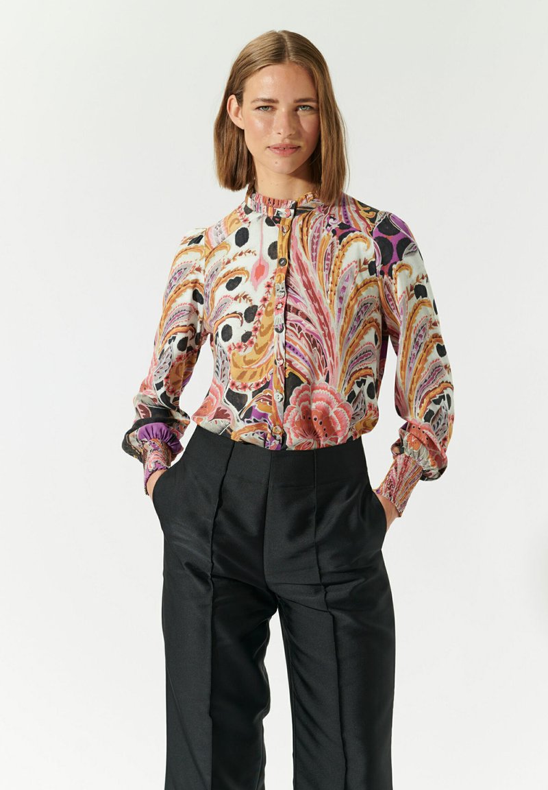 Blusa multicolore con motivo floreale e a spirale, con bottoni davanti, collo alto e maniche con polsini, abbinata a pantaloni neri a vita alta.