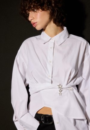 Femme aux cheveux bouclés portant une chemise blanche à boutons cintrée à la taille avec une épingle de sûreté argentée en forme d'étoile et un pantalon noir avec une ceinture.