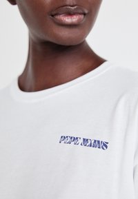 Balta kokvilnas T-kreklu ar zilu grafisku izdrukas "PEPE JEANS" kreisajā krūtī. Gluda tekstūra, apaļa izgriezuma kakla līnija, brīvs piegriezums.