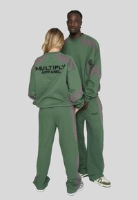 Două persoane îmbrăcate cu hanorace și pantaloni asortați, verzi și gri, cu sigla "Multiply Apparel", stând în fața unui fundal simplu.