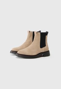 Botine din suede bej, cu panouri laterale elastice negre, vârf rotunjit și o talpă din cauciuc texturat cu un accent discret tricolor.