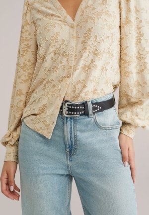 Béžová textúrovaná blúzka s dlhým rukávom zastrčená do svetlomodrých džínsov s čiernym opaskom so cvočkami a striebornou prackou, zobrazená od stredu trupu po stehná.