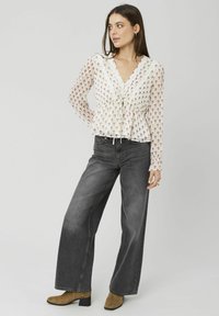 Femme portant un chemisier blanc à fleurs, un jean gris à taille haute et jambes larges, et des bottines marron, debout devant un fond uni.