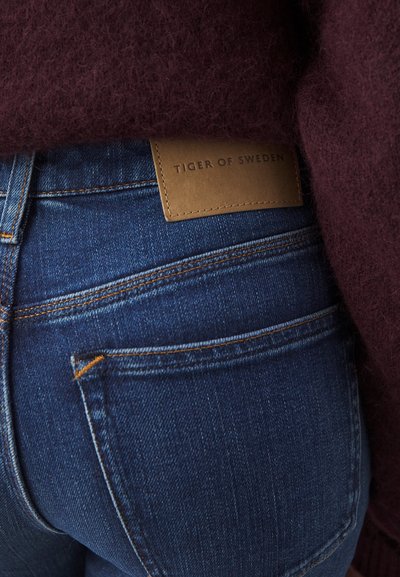 Vaqueros de mezclilla azul oscuro con una superficie texturizada, que presentan costuras naranjas y una etiqueta de cuero marrón con la inscripción "TIGER OF SWEDEN."