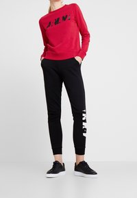 Felpa rossa con testo grafico nero e pantaloni jogger neri con stampa bianca sui lati, abbinati a sneakers nere. Tessuto in cotone, design aderente.
