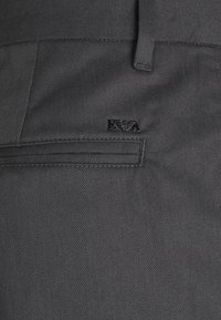 Šedé kalhoty z texturované látky s kapsou a malým černým logem. Výrazné jsou čisté linie a klasický střih.