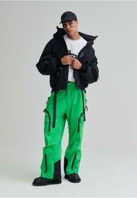 Chaqueta negra con capucha, camiseta blanca con gráfico, pantalones impermeables verdes brillantes con detalles y cremalleras negras, combinados con botas negras y gorra.