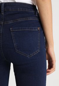 Jeans de mezclilla azul oscuro con una cintura alta, que presentan un bolsillo trasero con una forma distintiva y costuras marrones a lo largo de las costuras.