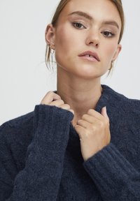 Pull bleu marine avec un motif texturé et un col montant à côtes. Il possède de longues manches avec de larges poignets côtelés. Matière douce.