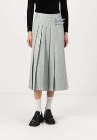 Jupe plissée gris clair à taille haute, ornée de deux boucles décoratives sur le côté. Associée à des chaussures noires épaisses et des chaussettes blanches.