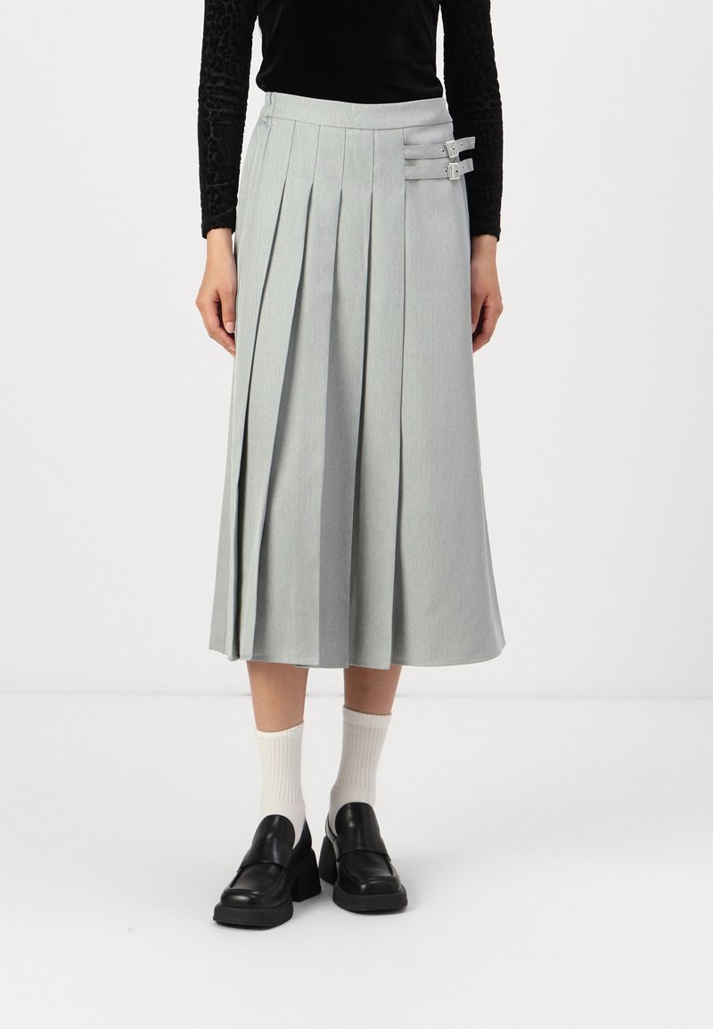 Jupe plissée gris clair à taille haute, ornée de deux boucles décoratives sur le côté. Associée à des chaussures noires épaisses et des chaussettes blanches.