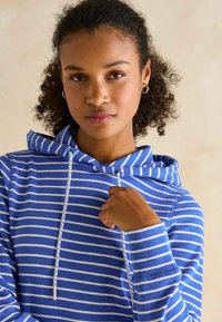 Žena s kučeravými vlasmi, ktorá má na sebe zlaté kruhové náušnice a modro-biele pruhované mikinu, drží šnúrku mikiny pri hrudníku.