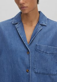 Denimshirt in medium blauw met een geknipte kraag, knoopsluiting aan de voorkant en borstzak. Zachte textuur met zichtbare stiksels.
