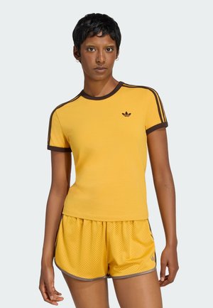 Persona che indossa una maglietta gialla Adidas con bordino marrone e pantaloncini a rete gialli abbinati, in piedi su uno sfondo chiaro.