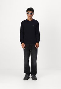 Σκούρο μπλε πλεκτό πουλόβερ με κανονική λαιμόκοψη, ριμπ μανσέτες και στρίφωμα; μαύρα παντελόνια σε χαλαρή γραμμή; μαύρα αθλητικά παπούτσια; ανακλαστικά γυαλιά ηλίου.