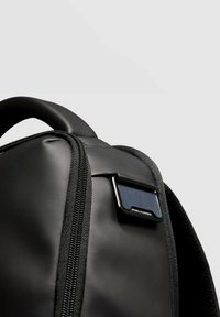 Sac à dos noir avec une finition mate et lisse. Dispose d'une fermeture éclair et d'une étiquette rectangulaire avec un accent métallique. Bretelles texturées.