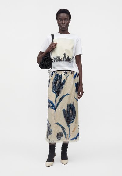 Femme portant un t-shirt graphique blanc, une jupe midi florale beige et bleue, des bottines chaussettes noires et tenant un sac à main noir sur fond blanc.