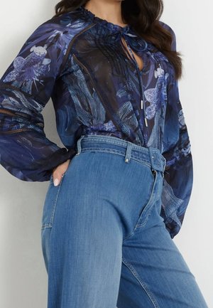 Kobieta w niebieskiej, prześwitującej, kwiatowej bluzce z długim rękawem, włożonej do jasnoniebieskich dżinsów z wysokim stanem i szerokim paskiem.