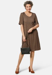 Kurzärmliges, braunes Kleid mit geometrischem Muster, lässiger Passform und gerafftem Taillenbereich. Kombiniert mit einer kleinen schwarzen Handtasche und braunen Absätzen.