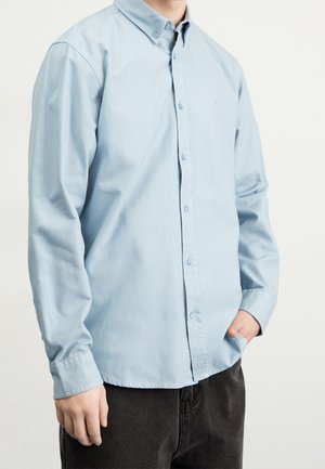 Mand iført en lyseblå skjorte med knapper og lange ærmer og mørke bukser, den ene hånd i lommen, mod en ensfarvet baggrund.