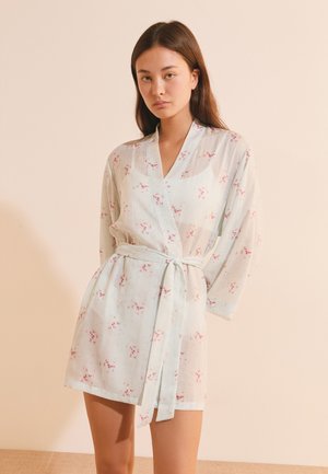 Jeune femme portant un peignoir léger à motif floral, ceinturé, manches longues, debout devant un fond beige.