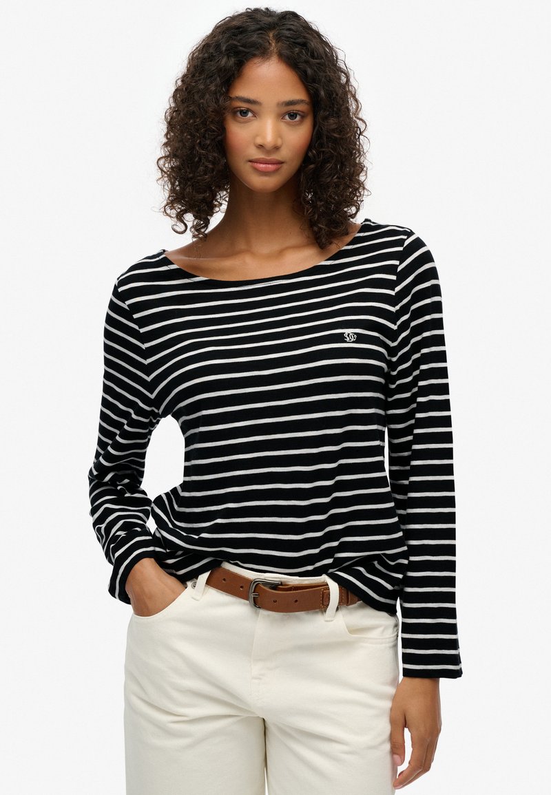 Camisa de rayas en blanco y negro de manga larga, con un escote amplio y tela texturizada. Pequeño detalle del logo en el pecho, combinada con pantalones beige.