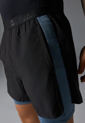Shorts de sport noirs avec doublure intérieure bleue et bande latérale, portés par une personne avec un bras et une main visibles sur un fond gris.