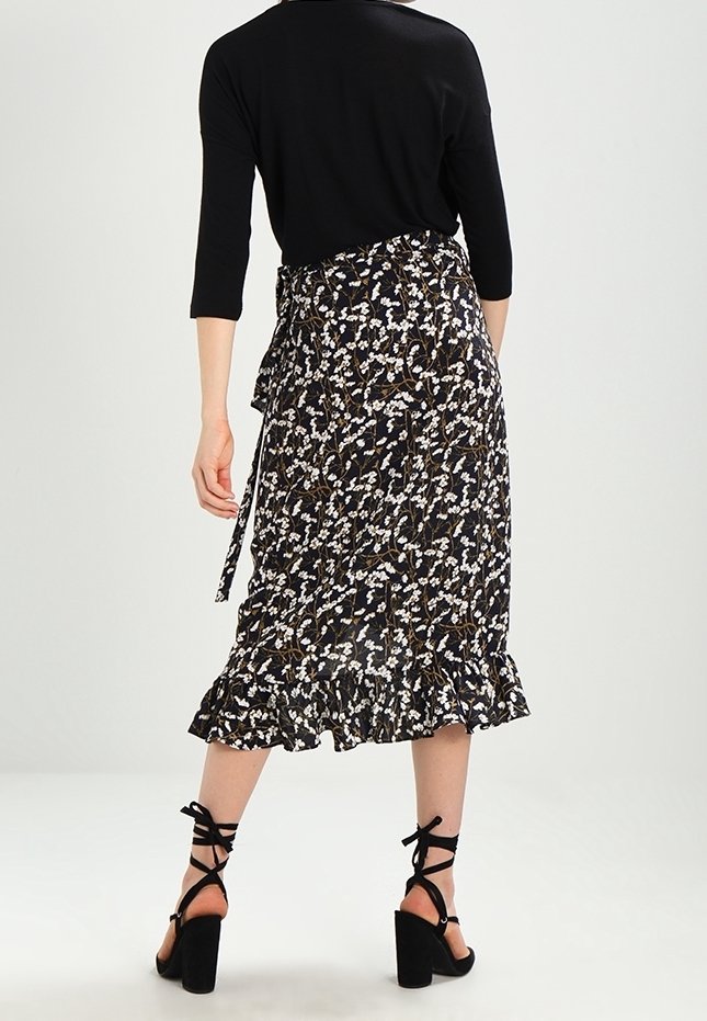 Femme portant une jupe midi noire à motifs floraux avec un ourlet à volants, un haut noir à manches trois-quarts et des chaussures à talons hauts noires à lacets, vue de dos.