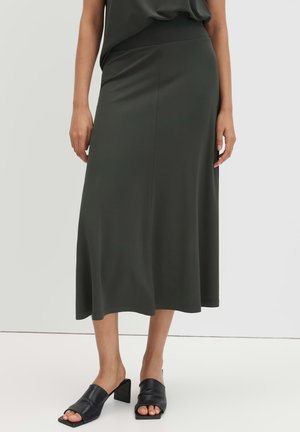 Femme portant une jupe midi vert foncé avec une taille ajustée, associée à des mules noires à talon bloc et à bout ouvert, se tenant devant un fond uni.