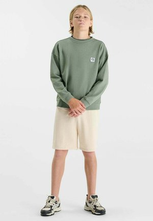 Tiener met blond haar staat met gevouwen handen, draagt een groene trui, beige korte broek en zwart-witte sneakers, op een witte achtergrond.