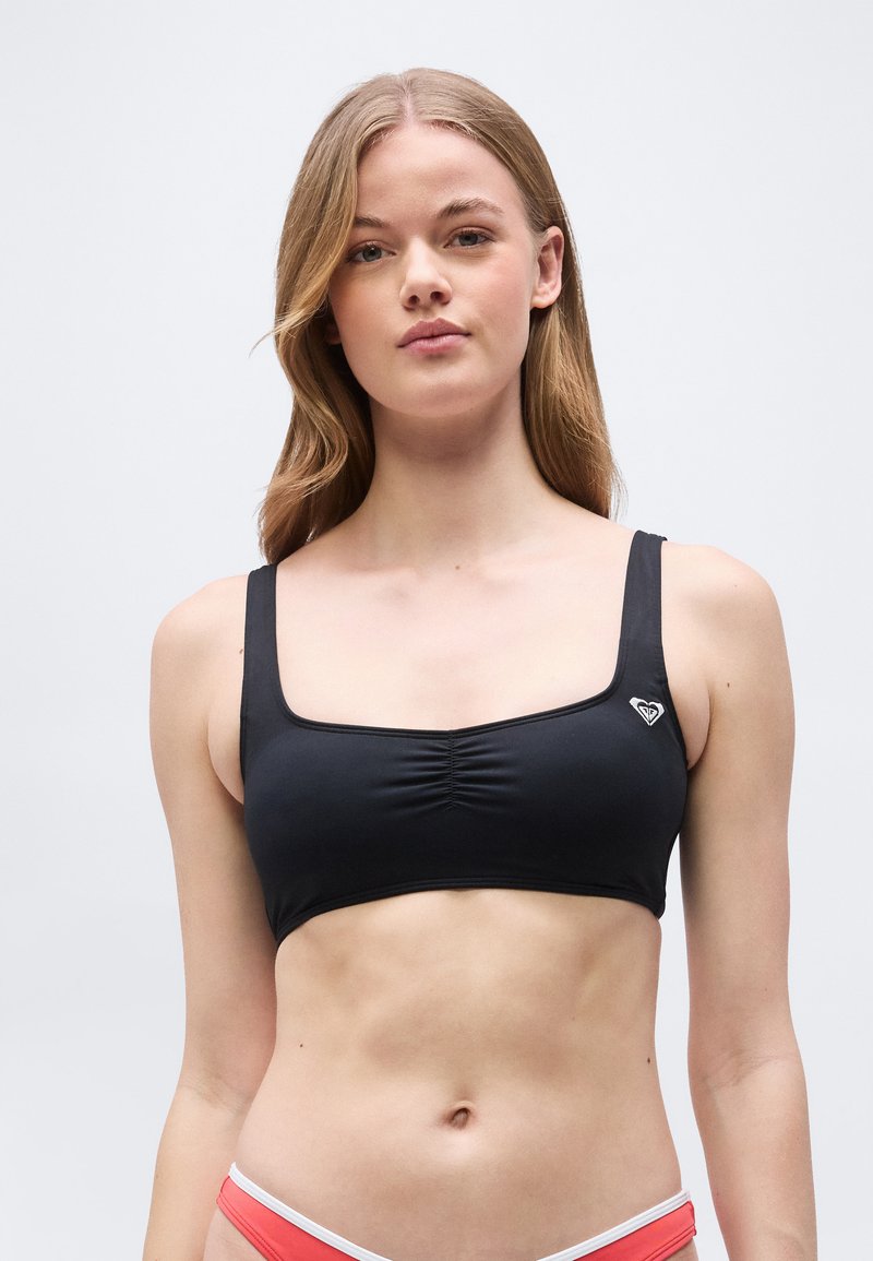 Νεαρή γυναίκα με μακριά ξανθά μαλλιά, φορώντας μαύρο κοντό τοπ χωρίς μανίκια και κόκκινα κάτω μέρη μπικίνι, με φόντο ένα απλό ανοιχτό χρώμα.