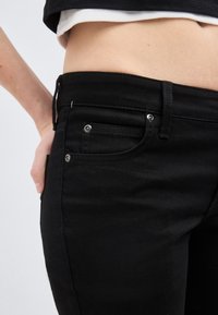 Κοντινό πλάνο της μέσης ενός ατόμου που φοράει μαύρα τζιν και μαύρο-άσπρο στρωματοποιημένο τοπ, με το ένα χέρι να στηρίζεται στο ισχίο του.