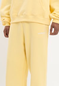 Sudadera amarilla con capucha y pantalones de chándal a juego, confeccionados con tela suave, que cuentan con un bolsillo tipo canguro y un logo bordado en la pierna.