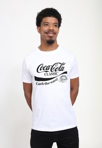Mand iført hvid t-shirt med sort "Coca-Cola Classic Catch the wave! Original Formula" logo, står med hænderne bag ryggen og smiler.