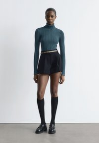 Frau trägt einen smaragdgrünen, gerippten Rollkragenpullover, schwarze, hochtaillierte Shorts mit Gürtel, schwarze kniehohe Socken und schwarze Slipper und steht vor einer weißen Wand.