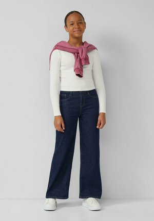 Brede denim jeans, donkerblauw, gecombineerd met een witte blouse met lange mouwen. Een roze sweater is om de schouders gestrikt, met witte sneakers.