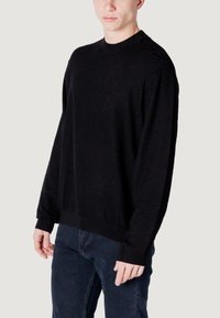 Maglione nero con un motivo testurizzato, design a girocollo e polsini a costine. Abbinato a jeans in denim scuro e uno sfondo neutro.