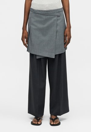 Ember réteges ruházatban: szürke aszimmetrikus szoknya széles fekete nadrág felett és fekete szandál, egyszerű háttér előtt áll.