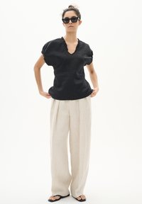 Zwarte linnen blouse met ruches bij de hals, gecombineerd met lichtbeige, wijdgevallen broeken. Het model draagt zwarte zonnnenbril en zwarte slippers.