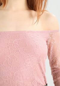 Top rosa scollato sulle spalle realizzato in pizzo, con motivi floreali e maniche lunghe in tessuto trasparente che si fondono in un tessuto solido in vita.