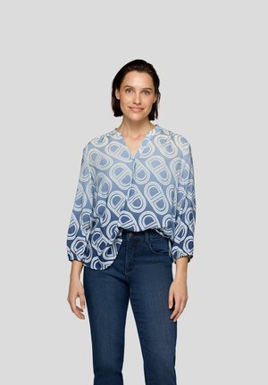 Knopfträger, hellblaue Bluse mit kreisförmigen Mustern in dunkleren Farbtönen, V-Ausschnitt und dreiviertel Ärmeln, kombiniert mit dunkelblauen Jeans.