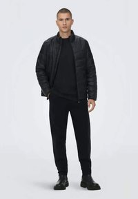 Sort pufferjakke med lynlås foran, parret med en sort crew neck skjorte og bukser. Sættet har et strømlinet, moderne design.