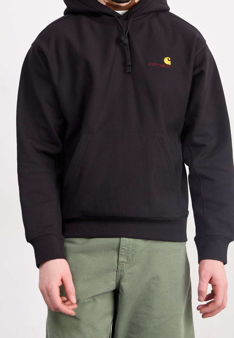 Schwarzer Baumwoll-Hoodie mit Fronttasche, Kapuze mit Kordelzug und kontrastierendem roten und gelben Logo auf der Brust. Modell trägt grüne Hosen.
