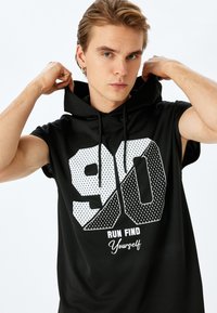 Zwarte mouwloze top met capuchon, voorzien van een groot wit "90"-grafiek met een stippenpatroon en de tekst "RUN FIND Yourself" eronder. Soepele textuur.