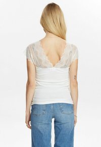 Top blanco de canalé con mangas cortas de encaje y espalda en V profunda. Estilizado con jeans azul, mostrando un diseño casual en capas y acentos texturizados.