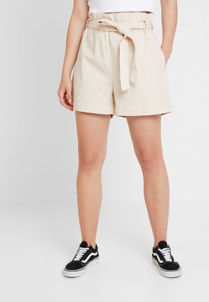Pantaloni corti in cotone beige con vita elastica e dettaglio a fiocco frontale, mostrati con sneakers nere dotate di lacci bianchi e design classico.